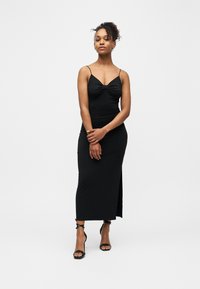 Femme debout portant une robe midi noire sans manches à fines bretelles et des sandales noires à talons hauts, sur un fond blanc uni.
