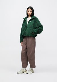 Πρ άσινο φλις μπουφάν με ψηλό γιακά, καφέ παντελόνι με κλασική φαρδιά γραμμή και πλαϊνά κουμπιά, και μπεζ αθλητικά παπούτσια με σκούρο λογότυπο σβούρας.