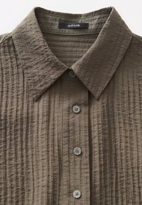 Gros plan d'une chemise marron texturée à boutons avec un col pointu et quatre boutons visibles à l'avant.