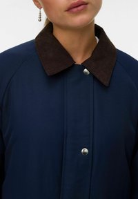 Giacca della marina con colletto in velluto a coste marrone, chiusura a scatto e una texture liscia. Presenta maniche raglan e dettagli minimi.
