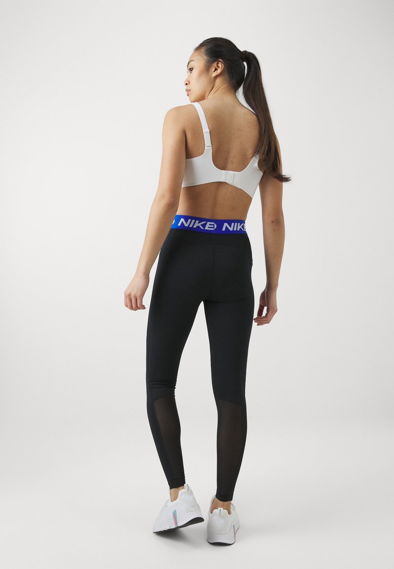 Soutien-gorge de sport blanc et leggings noirs avec des panneaux en maille au niveau des mollets. Les leggings présentent une ceinture bleue Nike. Le modèle fait face à l'arrière.