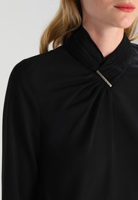 Svart blus med veckad halsringning och en silveraccent vid kragen. Tillverkad av mjukt tyg, den har en slank och figurnära design.