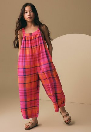 Jeune fille aux longs cheveux portant une combinaison ample à carreaux roses et orange sans manches, ainsi que des sandales beiges, se tenant devant un fond beige incurvé.