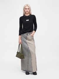 Femme aux longs cheveux blonds portant un haut noir à manches longues, une longue jupe en jean bicolore, tenant un sac à main vert, debout sur fond blanc.