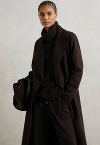 Abrigo largo de color marrón oscuro con un diseño a medida, que presenta un cuello con muescas y bolsillos laterales, combinado con un suéter de cuello alto oscuro y un bolso estructurado.