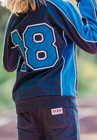 Sininen ja musta urheilutakki, jossa on suuri valkoisella ääriviivalla varustettu numero "18" selässä, sekä kontrastivalkoisia yksityiskohtia ja pehmeä kankaan tekstuuri.