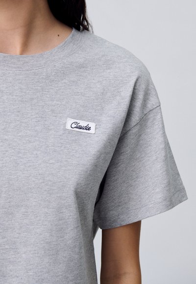 T-shirt in cotone grigio con maniche corte, dotato di una piccola etichetta bianca con il testo "Claude" in blu navy, posizionata sul petto.