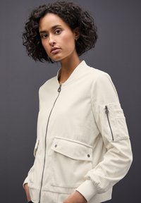 Veste bomber en velours côtelé de couleur claire avec une fermeture éclair, deux poches avant et des poignets côtelés. Présente une texture subtile et un design élancé.