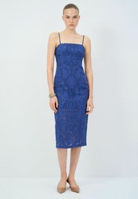 Robe midi en dentelle bleue avec des bretelles fines, mettant en avant un motif floral texturé, une coupe ajustée et un ourlet droit. Escarpins marron à bout pointu.