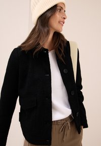 Donna che indossa un berretto a maglia color crema, un giubbotto nero con bottoni, una maglietta bianca e pantaloni beige con lacci, mentre porta una borsa a spalla beige chiaro.