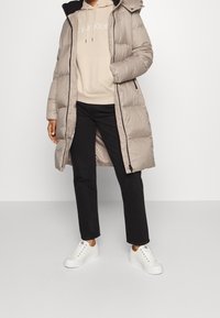 Giacca puffer beige con cappuccio, caratterizzata da un design trapuntato. Abbinata a un hoodie beige e pantaloni neri, completata da sneakers bianche.