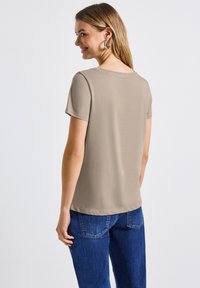 Camiseta beige de manga corta, con escote redondo, textura suave y un ajuste ligeramente holgado. Combinada con vaqueros azul oscuro, con detalles de costura visibles.