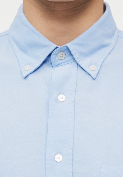 Nahaufnahme einer Person, die ein hellblaues Hemd mit drei sichtbaren weißen Knöpfen und einem geknöpften Kragen trägt.