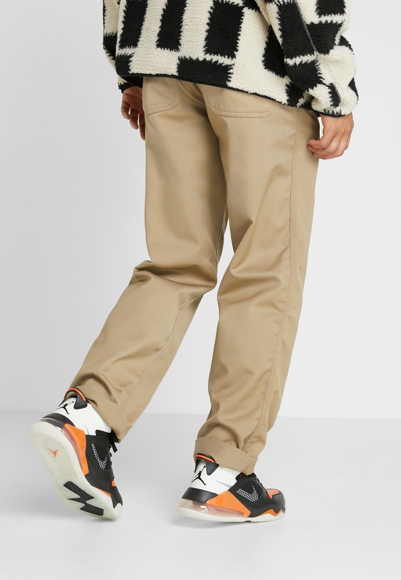 Khaki kalhoty s rovný fit, s bočními kapsami a hladkým povrchem, kombinované s černými a oranžovými teniskami na šedém pozadí.