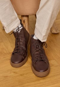 Brązowe skórzane buty typu high-top o gładkiej fakturze, z wyraźnymi sznurówkami i jasną gumową podeszwą. Noszone z białymi skarpetkami i beżowymi spodniami.