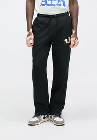 Čierne teplákové nohavice s nastaviteľným pásom na šnúrku, s zlatým logom "Nike" na ľavej nohe. Kombinované s svetlosivým tričkom s potlačou a teniskami.