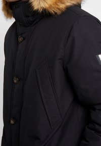 Sort vinterjakke med en blød hætte foret med pels. Har knaplukning og en sidelomme. Glat stof med mat finish.