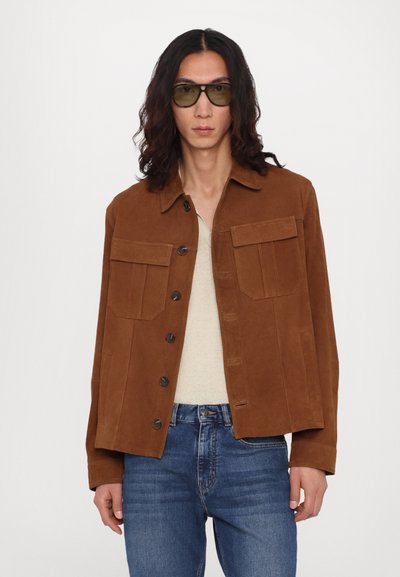 Veste en daim marron avec deux poches avant, fermeture par bouton et col pointu. Portée sur une chemise beige et associée à un jean bleu.