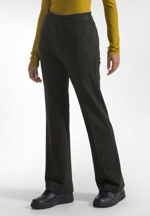 Persoon draagt zwarte wijde broek met naaldstrepen, zwarte schoenen en een mosterdkleurige top met lange mouwen, staand op een effen witte achtergrond.