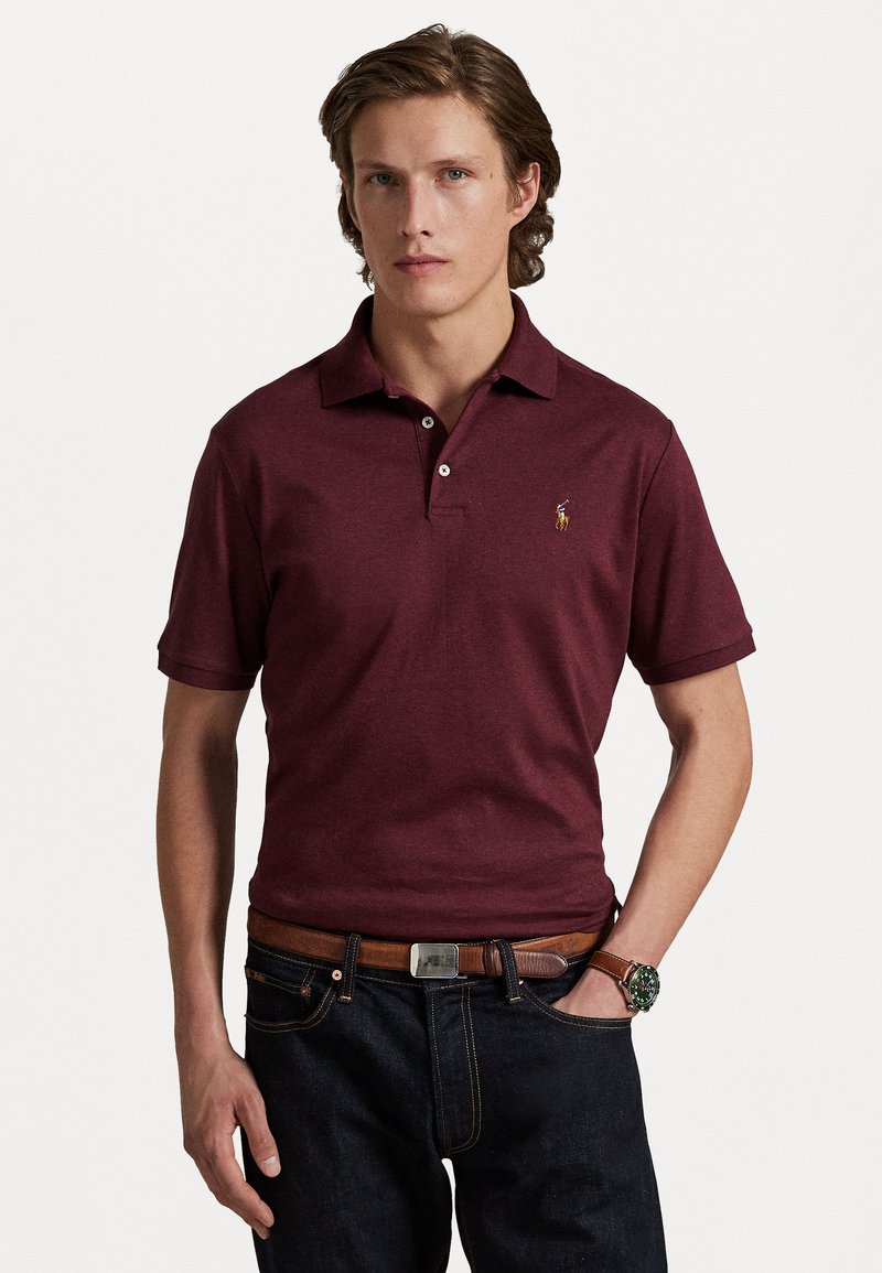 Tricou polo burgundy cu mânecă scurtă, cu un guler cu două butoane și logo brodat, asortat cu blugi închisi la culoare și o curea maro.