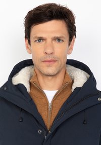 Hombre con pelo corto castaño que lleva una chaqueta con capucha de color navy con forro de felpa blanca y un suéter marrón con cremallera debajo.