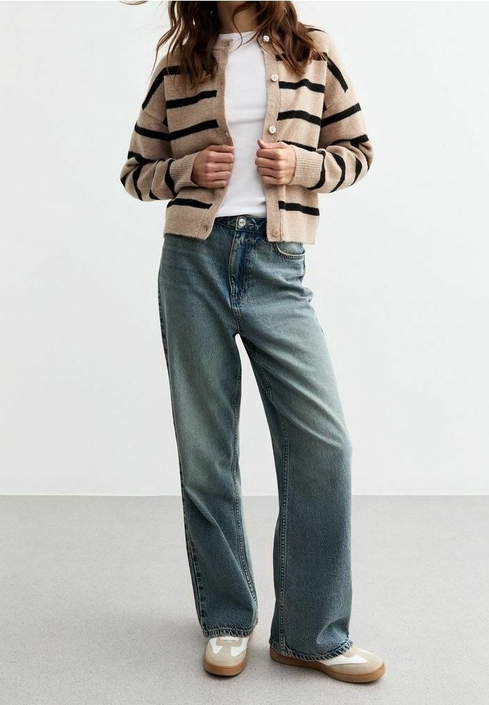 Femme portant un cardigan beige rayé par-dessus une chemise blanche, un jean bleu ample et des baskets beige-blanc, debout devant un fond blanc uni.