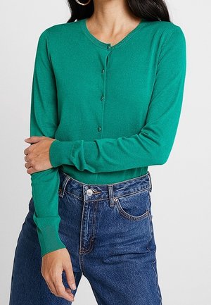 Kobieta ubrana w dopasowany zielony kardigan na guziki z długimi rękawami oraz niebieskie dżinsy z wysokim stanem na jednolitym tle.