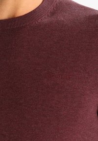 Burgunderfarbener Strickpullover mit Rundhalsausschnitt, der ein dezentes eingeprägtes Logo "ESPRIT" auf der Brust und eine glatte Textur aufweist.
