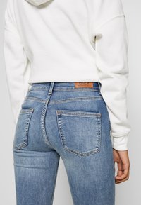 Persona que lleva pantalones vaqueros de mezclilla azul claro y una sudadera blanca, vista desde la parte posterior desde la cintura hasta la mitad del muslo.