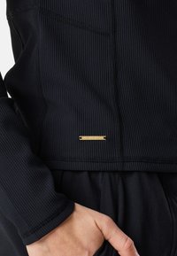 Svart ribbad tygjacka med en metallisk guldsymbol nära kanten, med en åtsittande design och dolda fickor på sidorna.