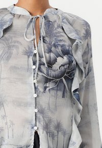 Bluză ușoară și transparentă în nuanțe de albastru, cu modele florale și de palmieri, guler f textured, decolteu legat și nasturi albi pe toată fața.