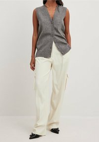 Grijze mouwloze vest met een diepe V-hals en vijf knopen, gecombineerd met witte wijde broek met zijvakjes en zwarte puntige schoenen.