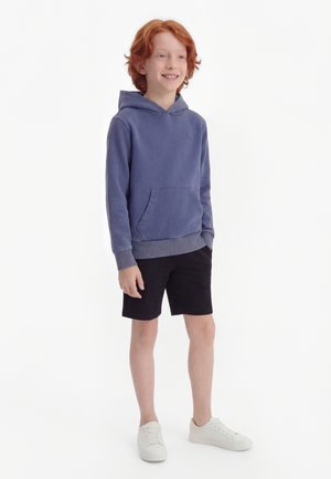 Smilende rødhåret dreng står iført en blå hættetrøje, sorte shorts og hvide sneakers mod en ensartet hvid baggrund.