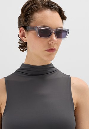 Donna con capelli corti e ricci indossa occhiali da sole rettangolari traslucidi e un top senza maniche grigio scuro con collo alto su sfondo semplice.