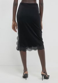 Schwarzer Samt-Midirock mit Spitze am Saum. Figurbetontes Design, glatte Textur und eine elegante Silhouette. Kombiniert mit schwarzen High-Heels.