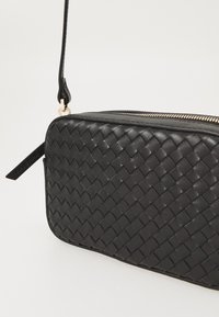 Borsa a pochette nera in pelle con texture intrecciata, hardware della cerniera dorato e tracolla staccabile. Forma rettangolare, design compatto.
