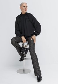 Schwarze Bluse mit gerafftem Ausschnitt, kombiniert mit strukturierten grauen Schlaghosen. Das Zubehör umfasst eine kleine schwarze Handtasche und Stiefeletten.