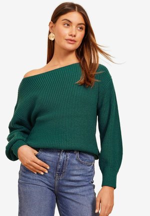 Grüner, gerippter Pullover mit einem schulterfreien Design, langen Ärmeln und strukturiertem Stoff. Kombiniert mit blauen Jeans.