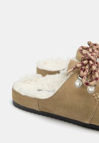 Bronasto rjavi čevlji iz zarezanega usnja brez vezalk z belo volneno oblogo, okrogle bež in vinsko barvne vezalke ter črnim gumijastim podplatom na belem ozadju.