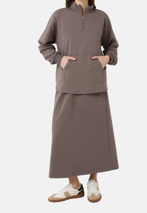 Personne portant un haut zippé taupe à manches longues avec poche avant et une jupe assortie longueur cheville, associée à des baskets blanches et beige.