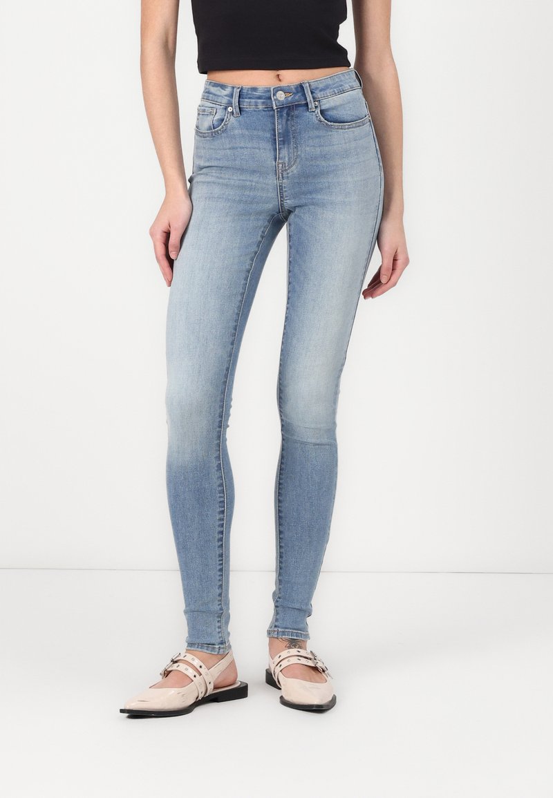 Jeans ajustados de color azul claro con cintura alta, lazos para cinturón y bolsillos frontales. Llevados con zapatos planos de color rosa pálido que presentan un diseño de tiras.