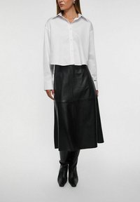 Hvid cropped skjorte med knapper og spredt krave, kombineret med en lang sort lædernederdel og knæhøje sorte støvler. Glatte teksturer.
