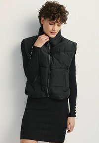 Czarna kamizelka puchowa z wysokim kołnierzem i zapięciem na zamek z przodu, wyposażona w dwie boczne kieszenie. Nosi ją się nad teksturowaną czarną sukienką z guzikami.