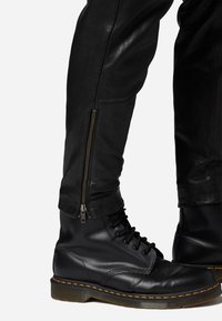 Zwarte leren gevechtslaarzen met vetersluiting, ronde neus en gele stiksels. Slanke zwarte leren broek met ritsdetail bij de enkels.