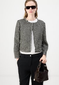 Jachetă hârtie cu model houndstooth alb-negru, cu un design scurt, detalii cu franjuri, purtată peste un top alb. Modelul ține o geantă texturată de culoare maro închis.