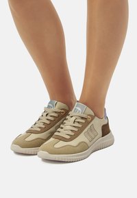 Zapatillas beige y marrón con materiales de ante y malla, con una suela texturizada, diseño de cordones y un logo en el lateral.