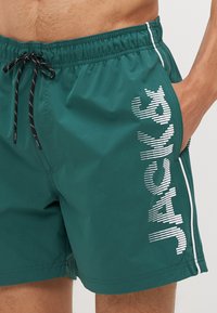 Teal színű fürdőshorts, könnyű anyagból készült. Fekete húzózsinórral, fehér oldalsávval és hullámos formájú grafikai szöveggel rendelkezik.