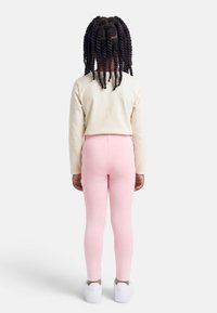 Παιδί με στριφογυριστές πλεξούδες στέκεται γυρισμένο από την πλάτη, φοράει κρεμ μακρυμάνικη μπλούζα, ροζ κολάν και λευκά αθλητικά σε απλό φόντο.