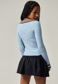 Hellblauer gerippter Pullover mit langen Ärmeln, versehen mit einem braun-weißen gestreiften Kragen. Kombiniert mit einem schwarzen Faltenrock mit weißen Streifen.