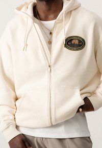 Roomkleurige hoodie met ritssluiting, een voorvak, trekkoorden en een geborduurd boomlogo met het jaartal 2018. Draagt een witte geribbelde shirt eronder.